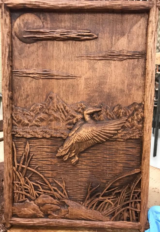 Wooden carved panel of a flying bird over marsh reeds, in warm brown tones.