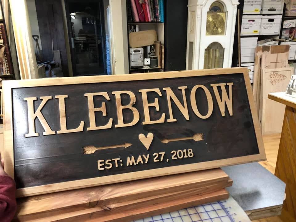 Wooden sign reading “KLEBENON” with “EST: MAY 27, 2018” on a workshop bench