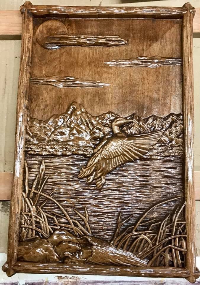 Brown carved wall plaque of a duck flying over a lake with mountains and reeds