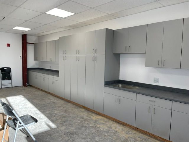 Empty community room with gray storage cabinets, counters, and folding chairs under fluorescent lights.