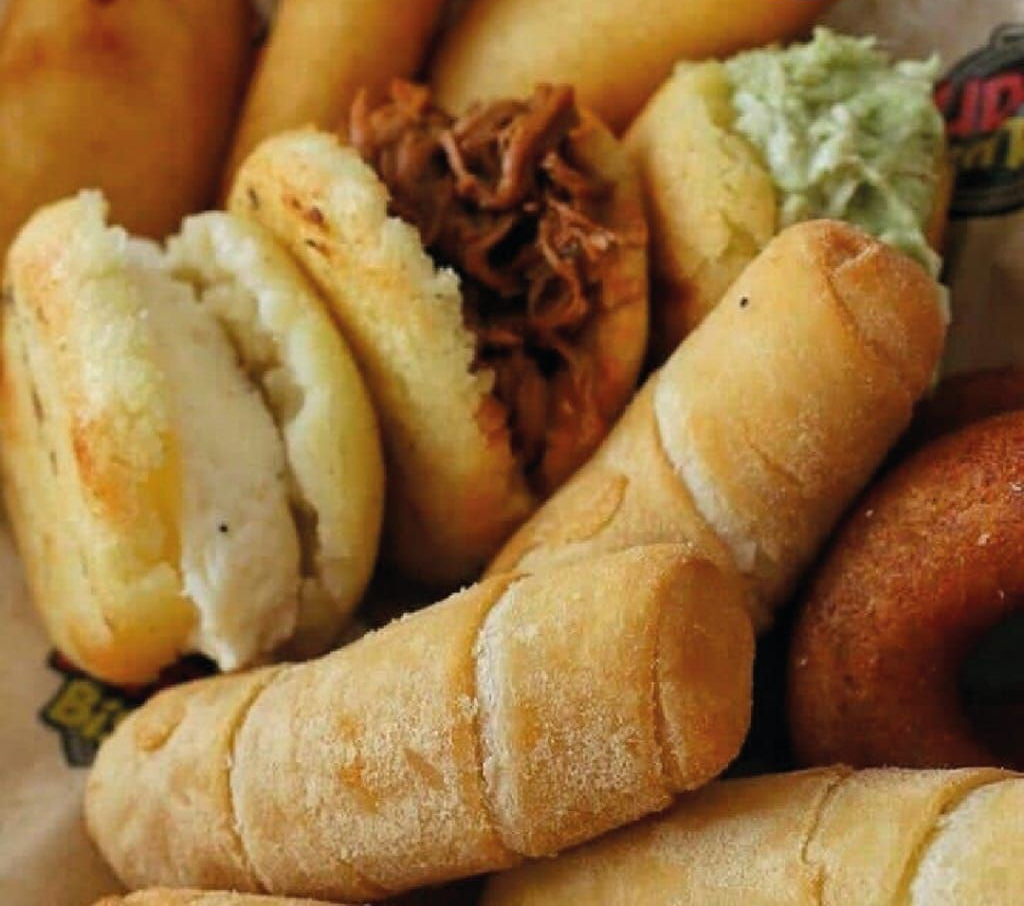 Primer plano de varios pasteles sudamericanos: arepas, tequeños y otros, algunos rellenos, en una canasta.