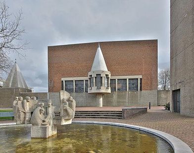 Een ronde fontein met een centraal beeldhouwwerk staat voor een bakstenen gebouw met een opvallende, puntige witte structuur.