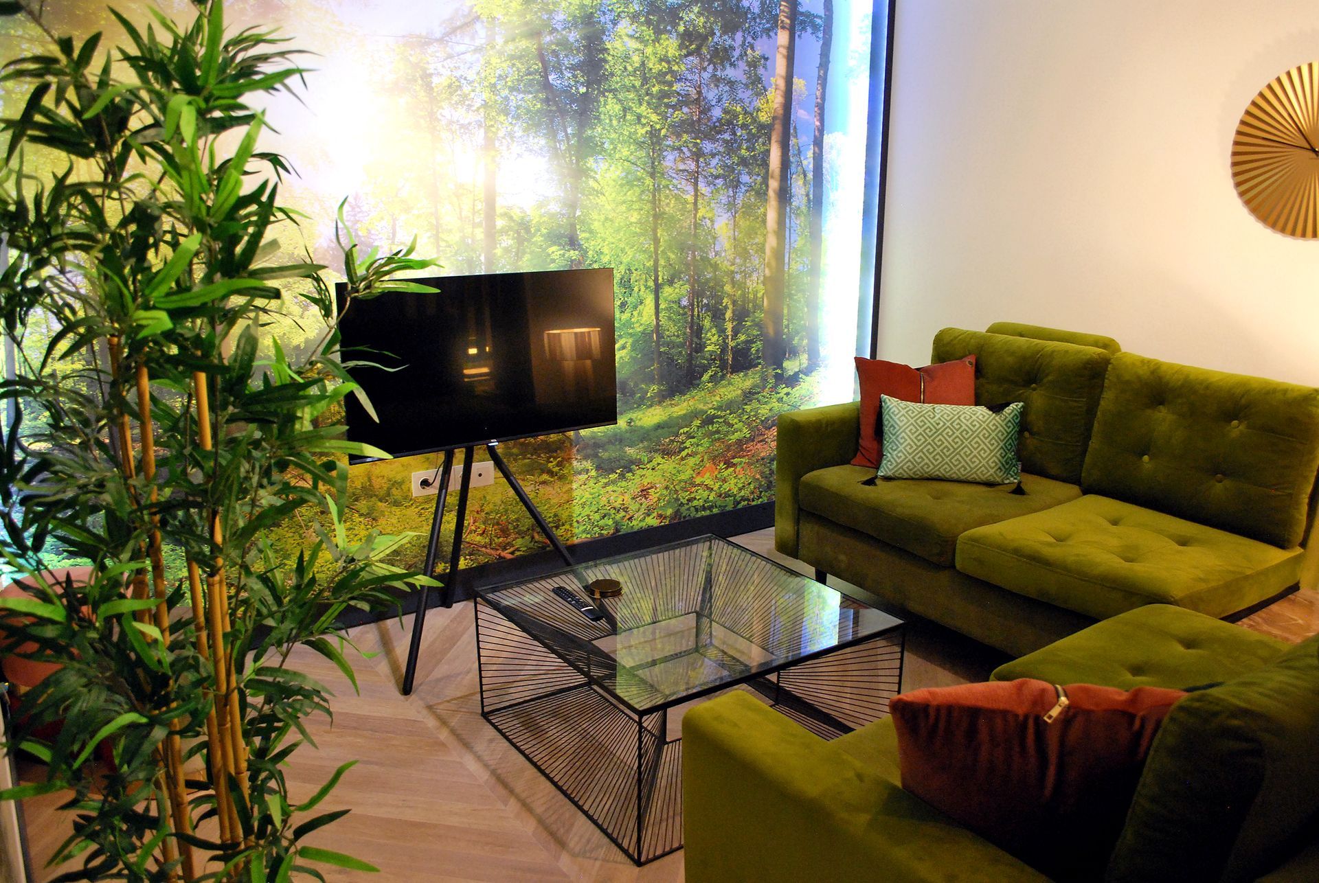 Een knusse lounge met groene banken, een tafel met glazen blad, een bamboeplant en een grote muurschildering van een zonovergoten bos.