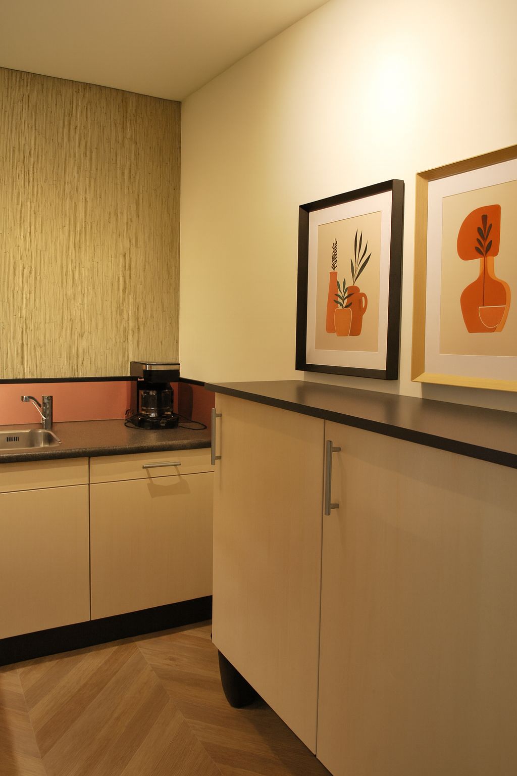 Een kleine kitchenette met lichtgekleurde kastjes, een spoelbak, een koffiezetapparaat en twee ingelijste abstracte kunstwerken aan de muur.