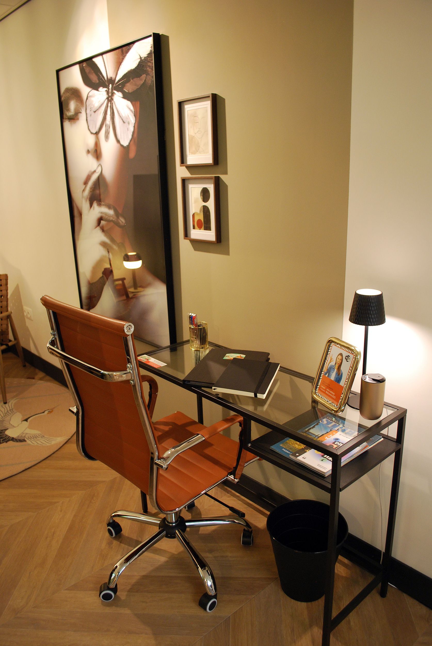 Een moderne werkplek met een beige leren bureaustoel, een bureau met glazen blad, een kleine lamp, wanddecoratie en een zwarte prullenbak.