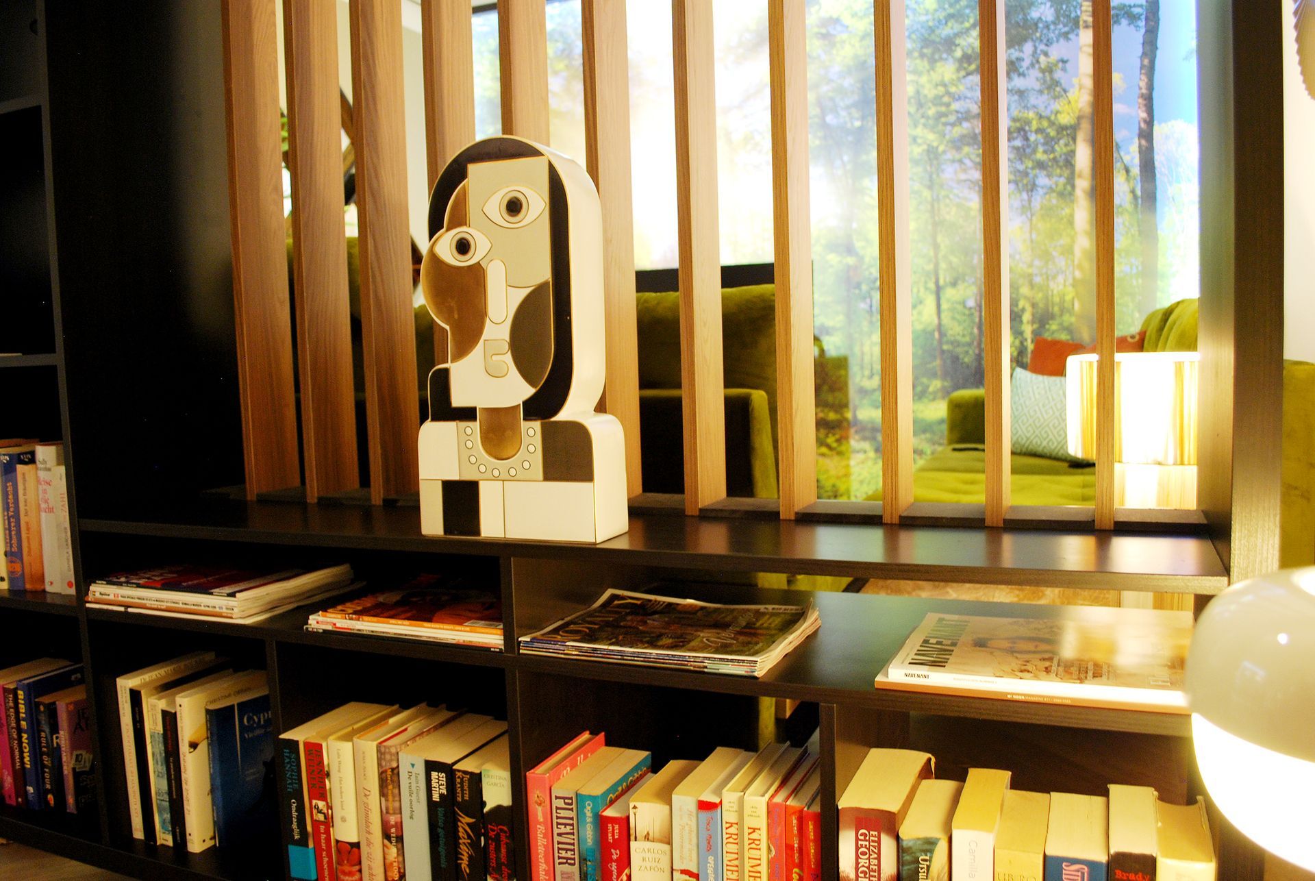 Een minimalistische abstracte sculptuur op een zwarte boekenplank gevuld met diverse boeken, zichtbaar door verticale houten latten.