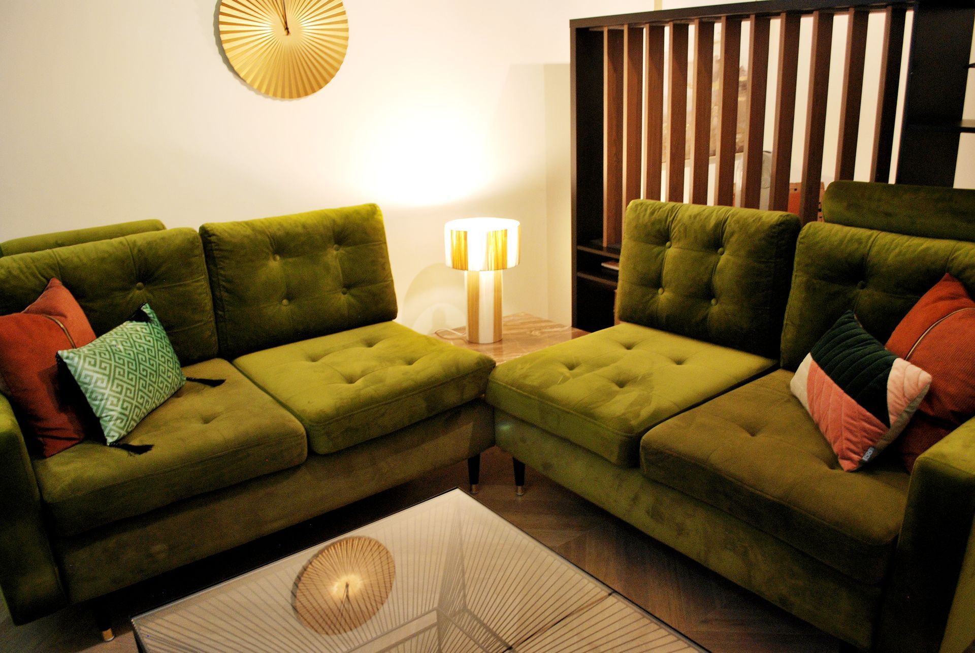 In een woonkamer staan ​​twee groene, gecapitonneerde banken tegenover elkaar, met kleurrijke sierkussens, een salontafel en een sfeervolle lamp.