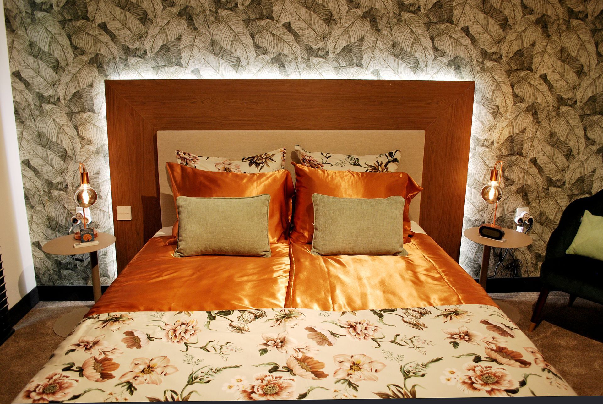Een knusse slaapkamer is voorzien van een bed met oranje beddengoed, kussens met bloemenpatroon, een houten hoofdbord en sfeervolle wandverlichting.