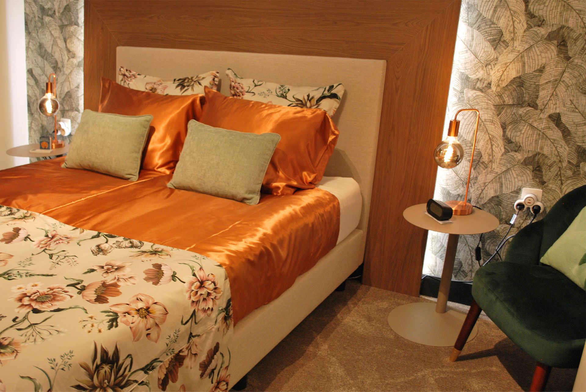 Een bed met oranje satijnen beddengoed, een bloemenplaid en lichtgroene kussens in een kamer met behang met een structuur en patroon.