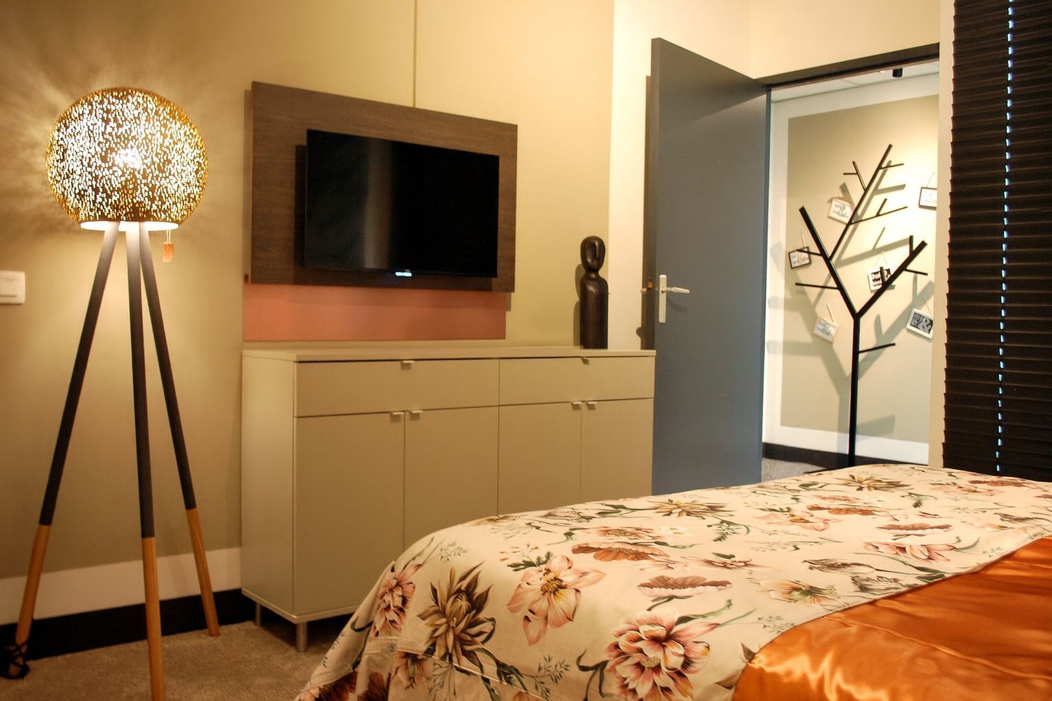 Een moderne slaapkamer met een staande lamp op een statief, een televisie boven een grijs kastje en een bed met bloemenpatroon.