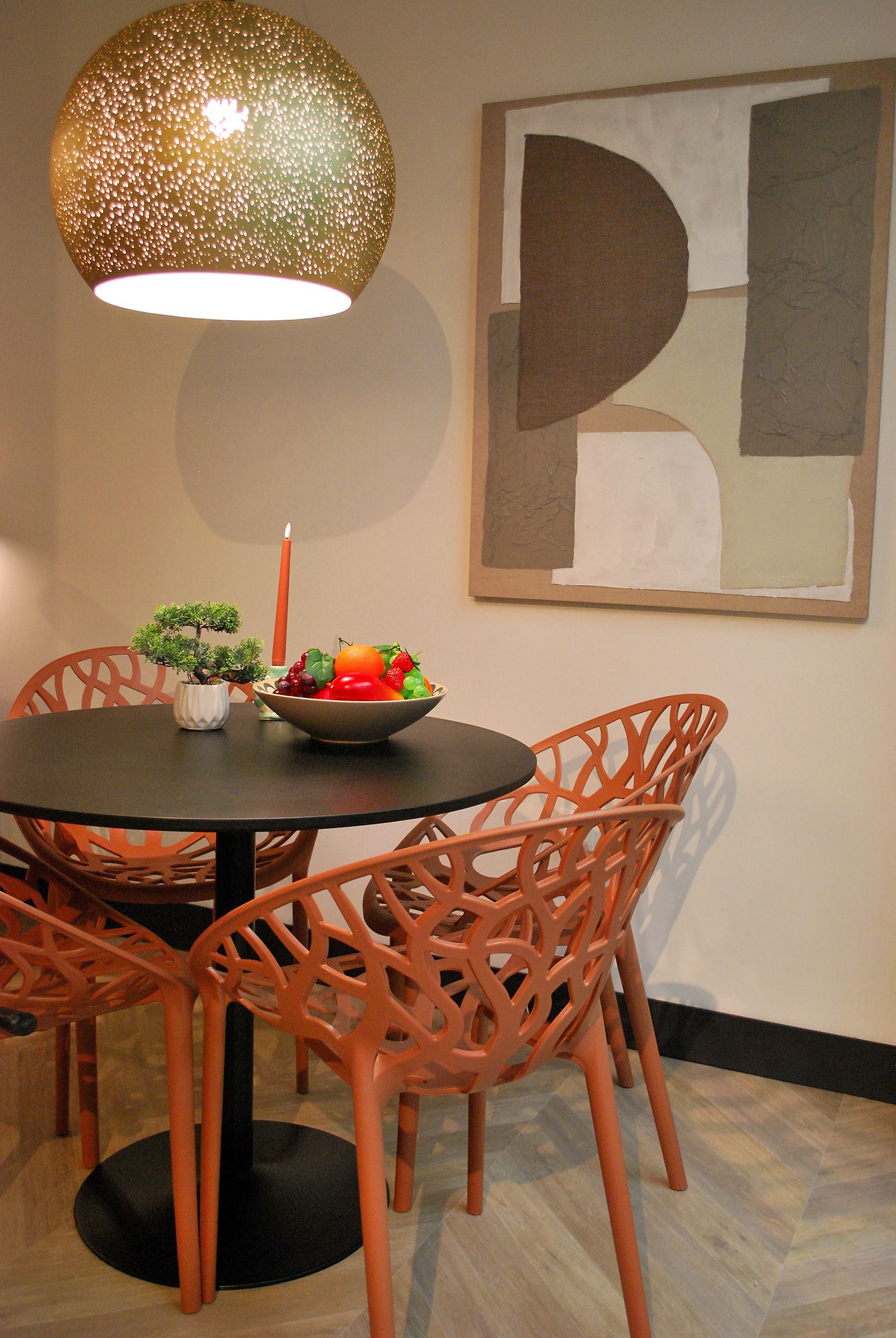 Een kleine ronde zwarte tafel met vier oranje stoelen met patroon, een hanglamp en geometrische muurdecoratie in een moderne kamer.