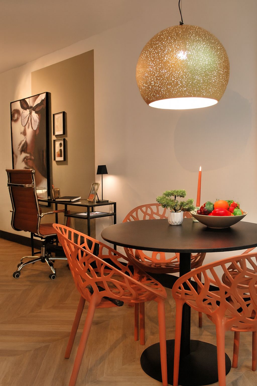 Een eethoek met een donkere ronde tafel, oranje stoelen met een rasterpatroon, een gouden hanglamp en een kleine werkplek erachter.