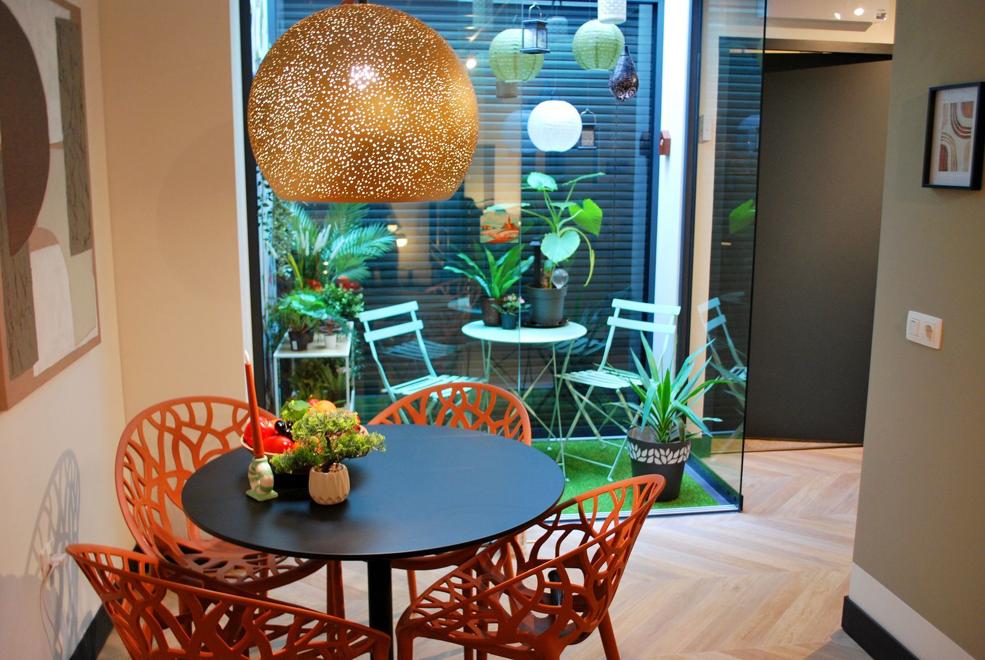 Een ronde eettafel met vier oranje, lasergesneden stoelen onder een decoratieve hanglamp, met uitzicht op een glazen patio.