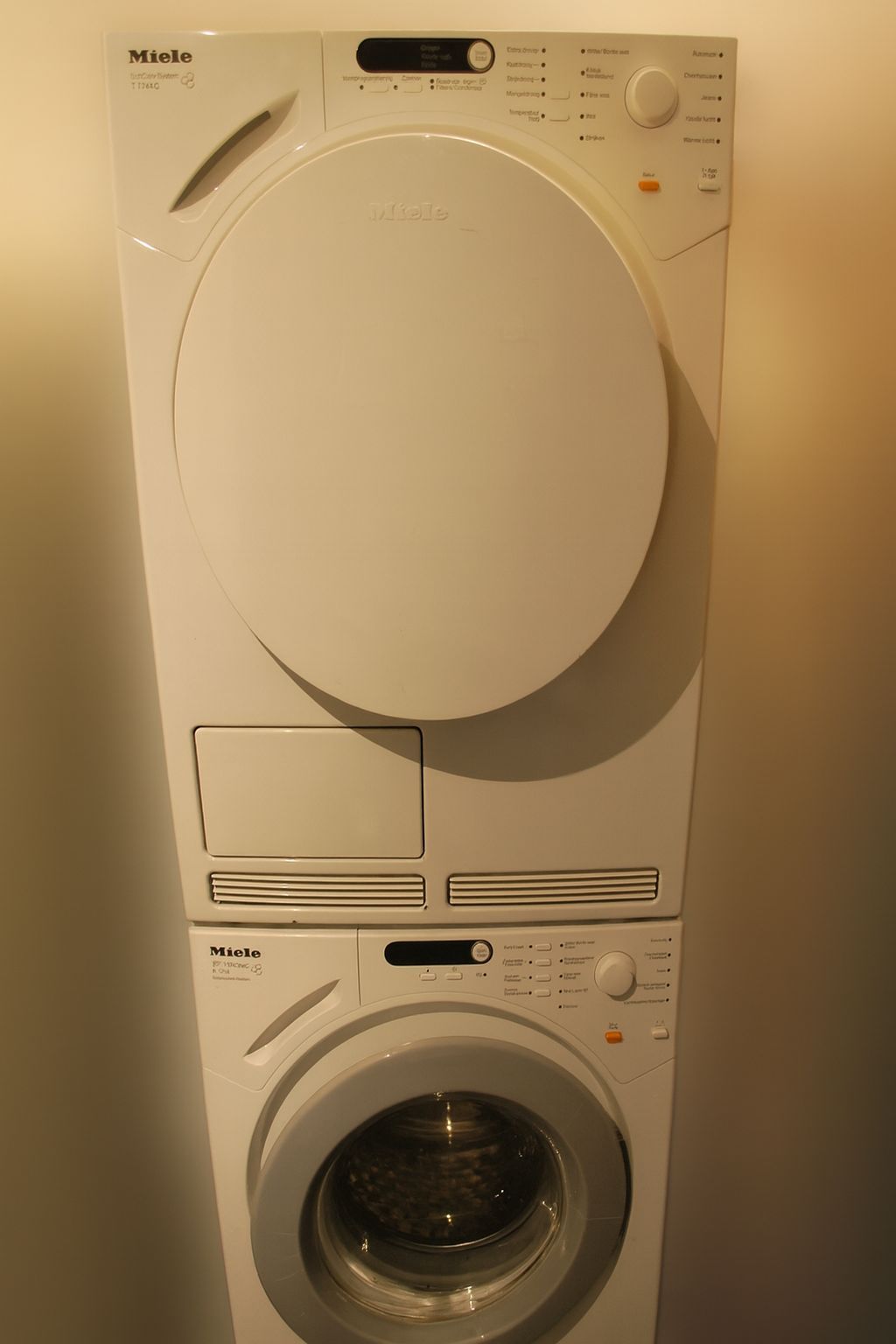 Een witte wasdroger van het merk Miele, bovenop een bijpassende wasmachine met voorlader, tegen een effen muur.