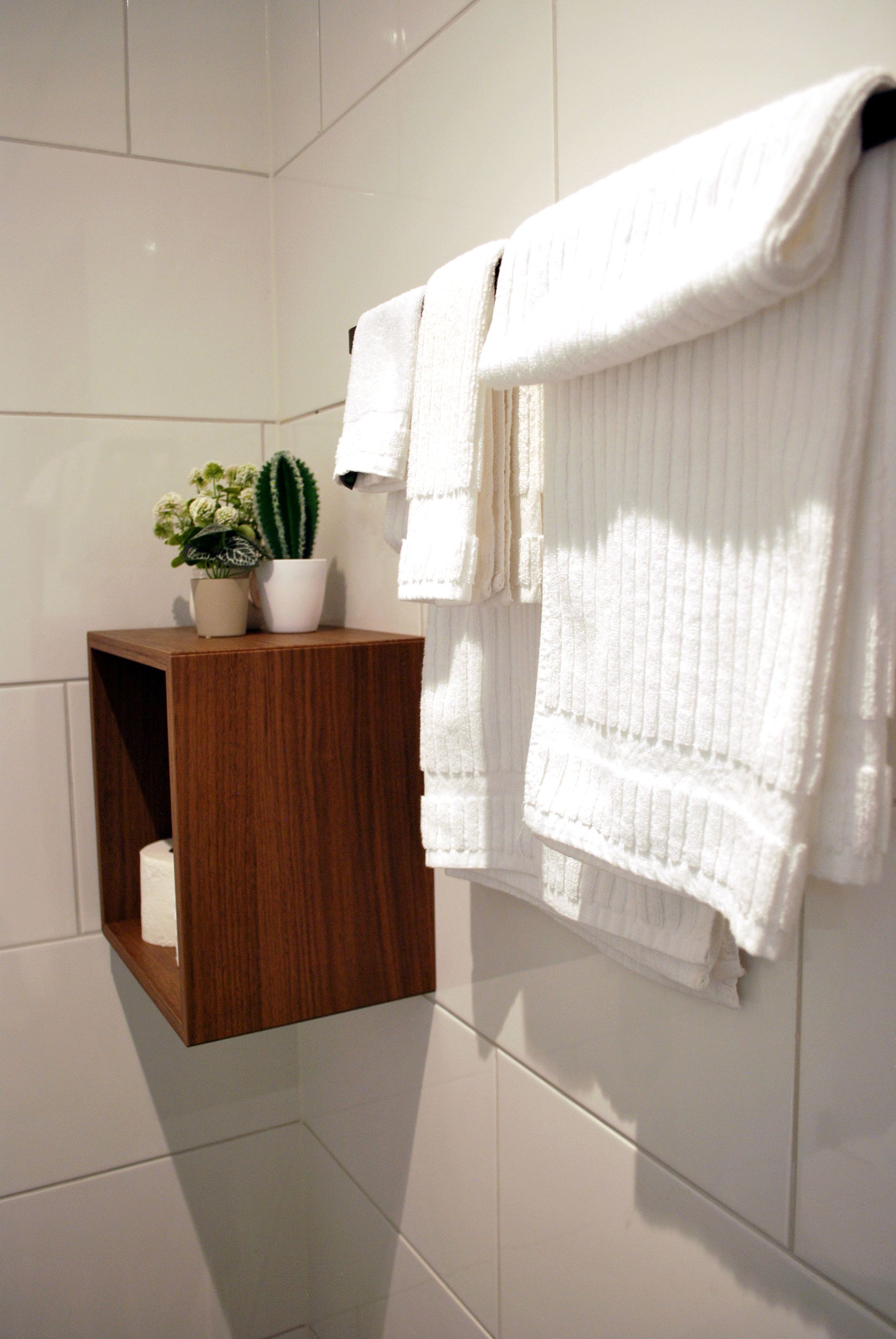 Witte handdoeken hangen aan een rek naast een klein houten wandplankje waarop twee potplanten en een rol toiletpapier staan.