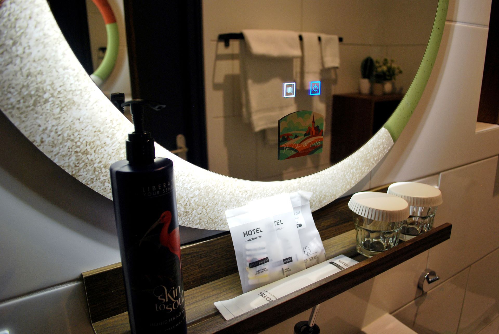 Een badkamermeubel met een ronde spiegel met ingebouwde digitale pictogrammen, een lotionfles, toiletartikelen en twee glazen.