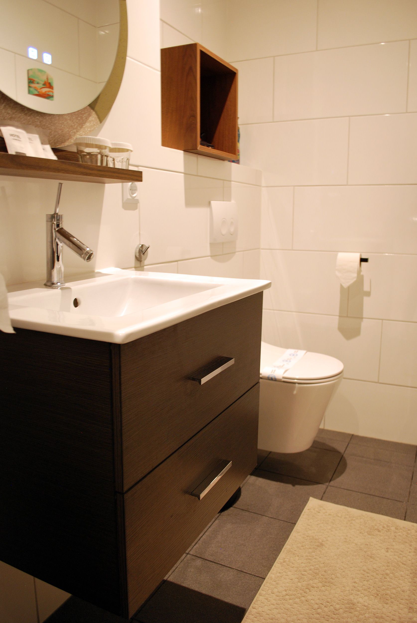 Een moderne hotelbadkamer met een witte wastafel op een donkerhouten badmeubel, een ronde spiegel en een aan de muur gemonteerd toilet.