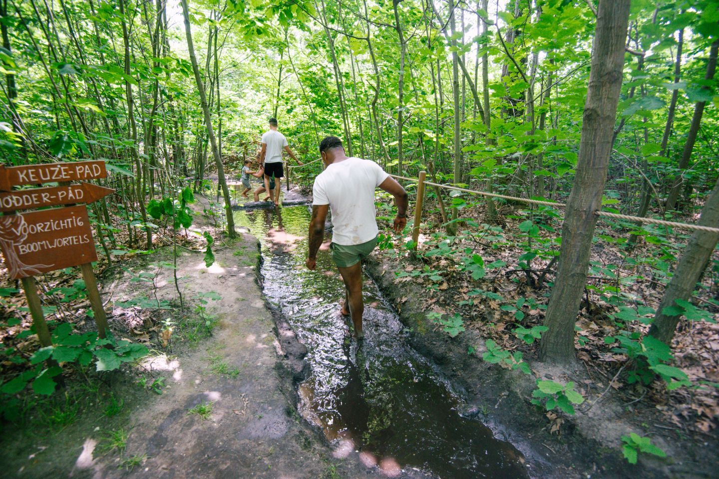 Twee mensen lopen door een ondiepe, modderige greppel in een weelderig groen bos, met een houten bord aan de linkerkant.