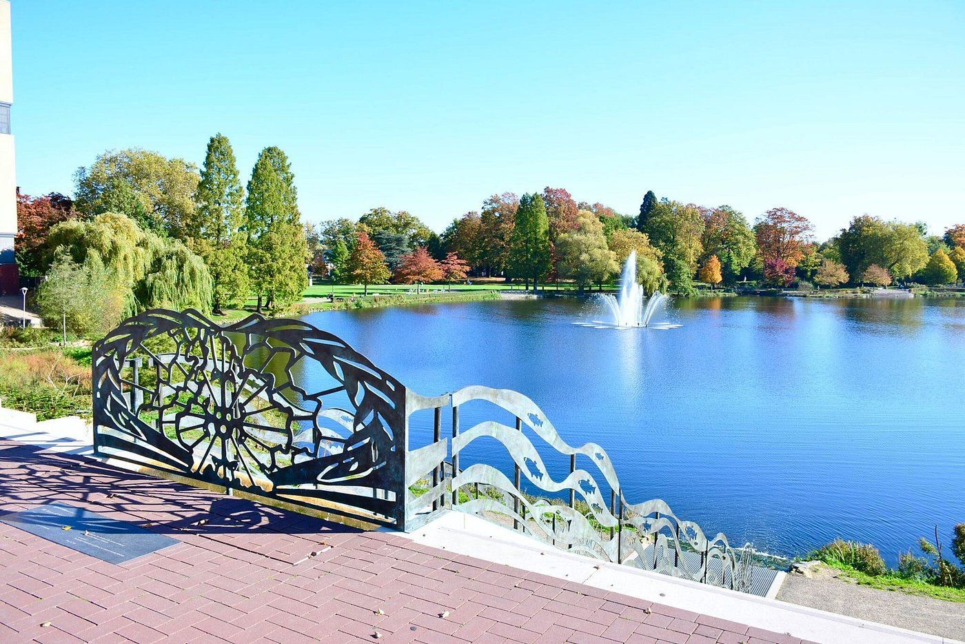 Decoratieve metalen balustrade met uitzicht op een meer met een fontein en herfstbomen onder een helderblauwe hemel.