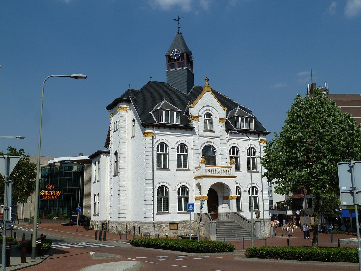Een historisch wit gebouw met een donker dak en een klokkentoren staat op een stadsplein onder een helderblauwe hemel.