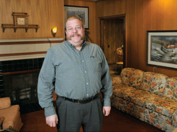 Man with mustache stands in front of a fireplace and floral couch in a wood-paneled room.