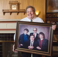 Man holding framed family portrait, smiling. Portrait shows four people, formal wear. Fireplace in background.