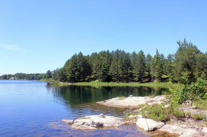 Lakeshore with clear blue water reflecting trees and a bright, cloudless sky.