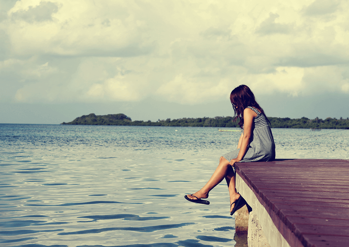 Woman sitting on a wooden dock, feet dangling over water, looking out at the ocean on a cloudy day.