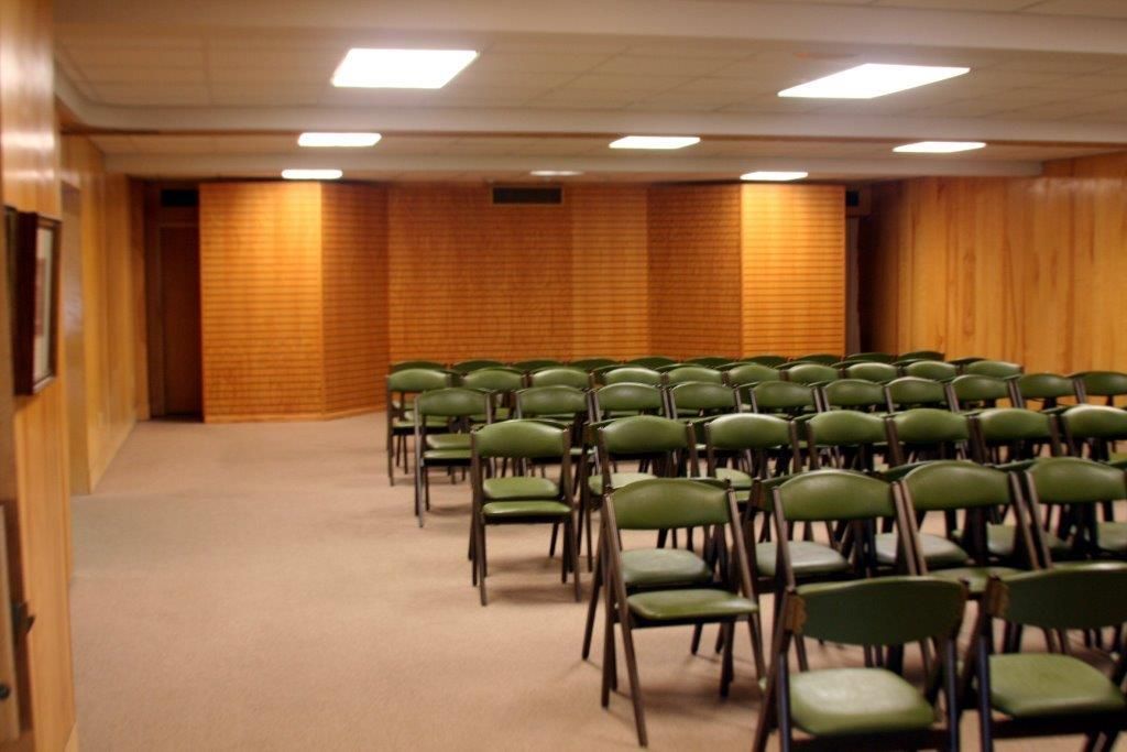 Rows of green chairs face a wood-paneled wall and ceiling in a beige-carpeted room.