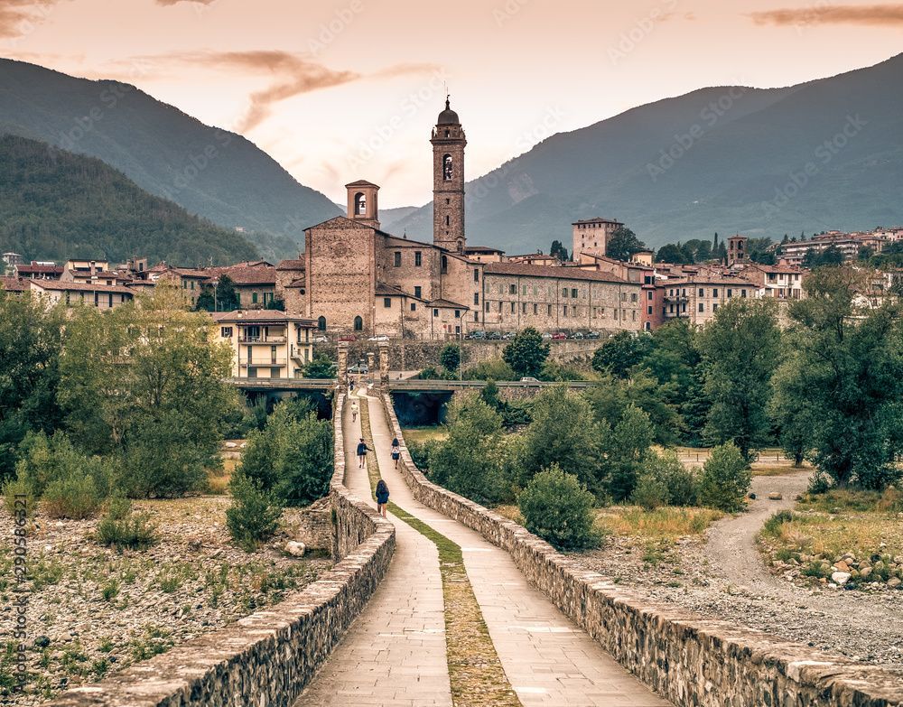 Stone bridge leads to medieval town with mountains in the background.