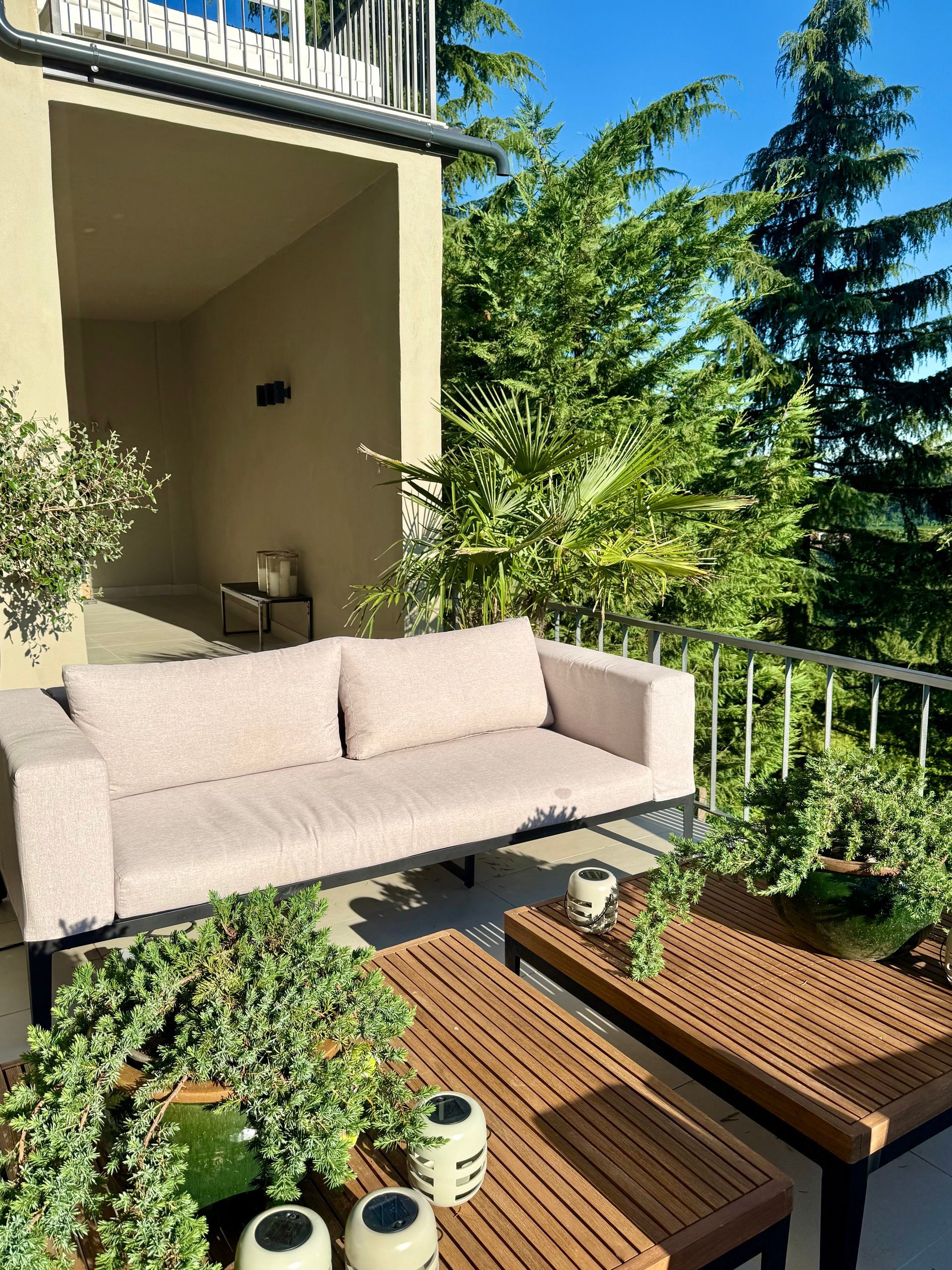 Outdoor patio with a light-colored sofa, two wooden tables, and lush greenery.