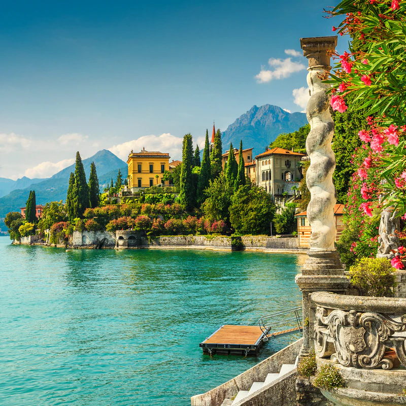 Lake Como, Italy. Buildings on shore with mountains in the distance, turquoise water, flowers in foreground.