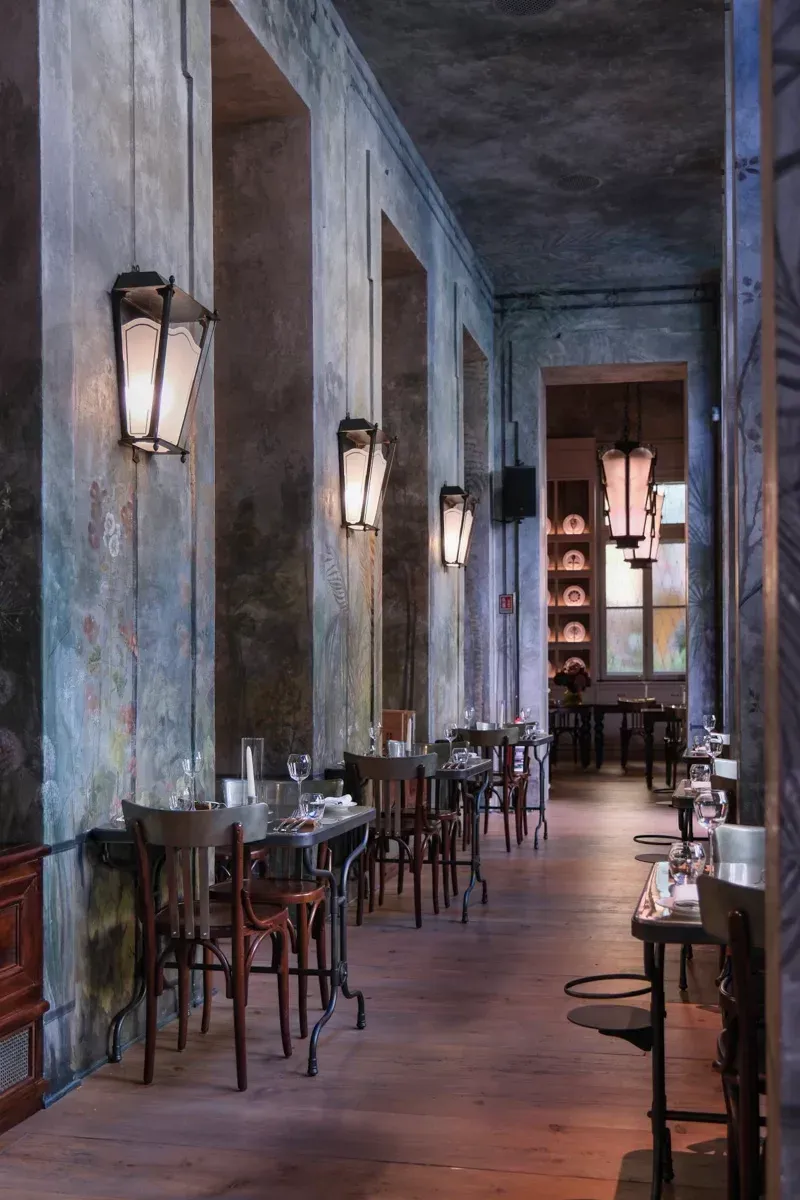 Long, narrow restaurant with tables, chairs, sconce lighting, and weathered gray walls; warm-toned floor.
