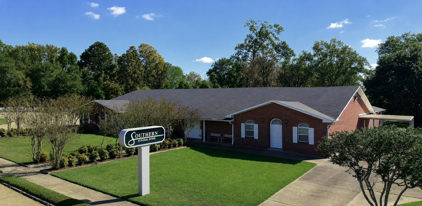 A brick building with a dark roof and a sign in front, surrounded by a green lawn and trees.