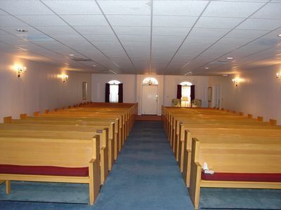 Interior of a chapel with rows of wooden pews, blue carpet, and a central aisle leading to a door.