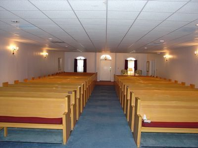 Empty church sanctuary with wooden pews, blue carpet, and arched windows centered on the aisle