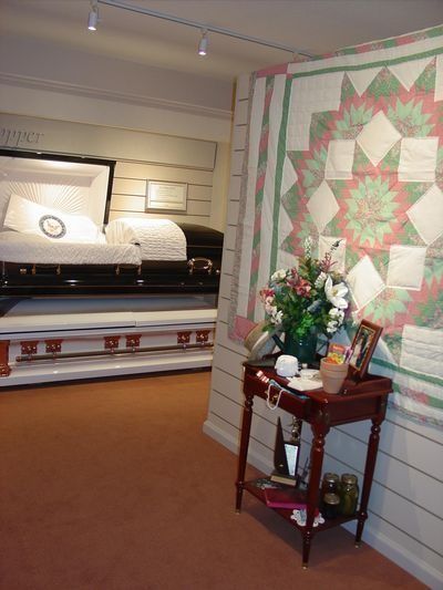 Funeral home interior with open caskets, small table with flowers and a quilt on the wall.