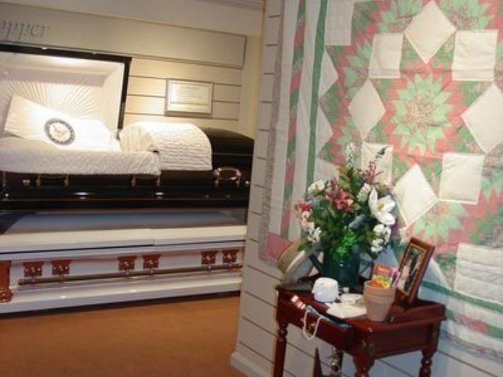 Interior of a funeral home with an open casket, flowers, and a quilt on the wall.