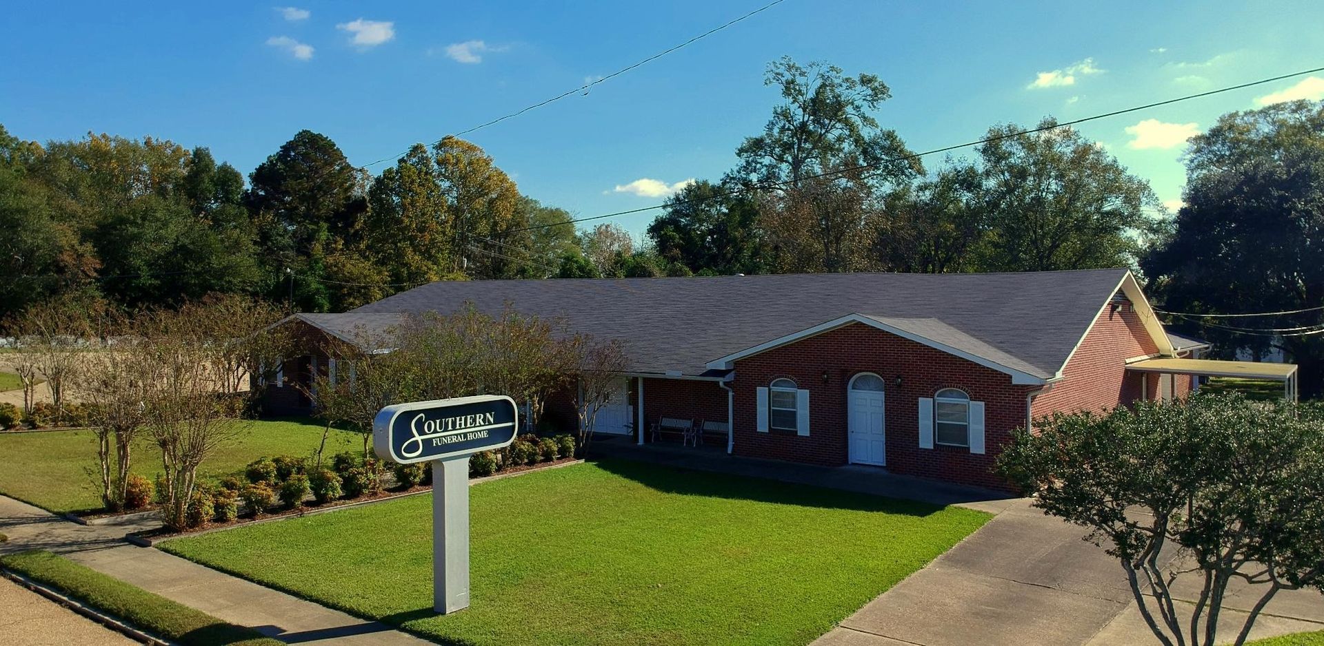 A brick building with a dark roof and a sign in front, surrounded by trees and a green lawn under a blue sky.