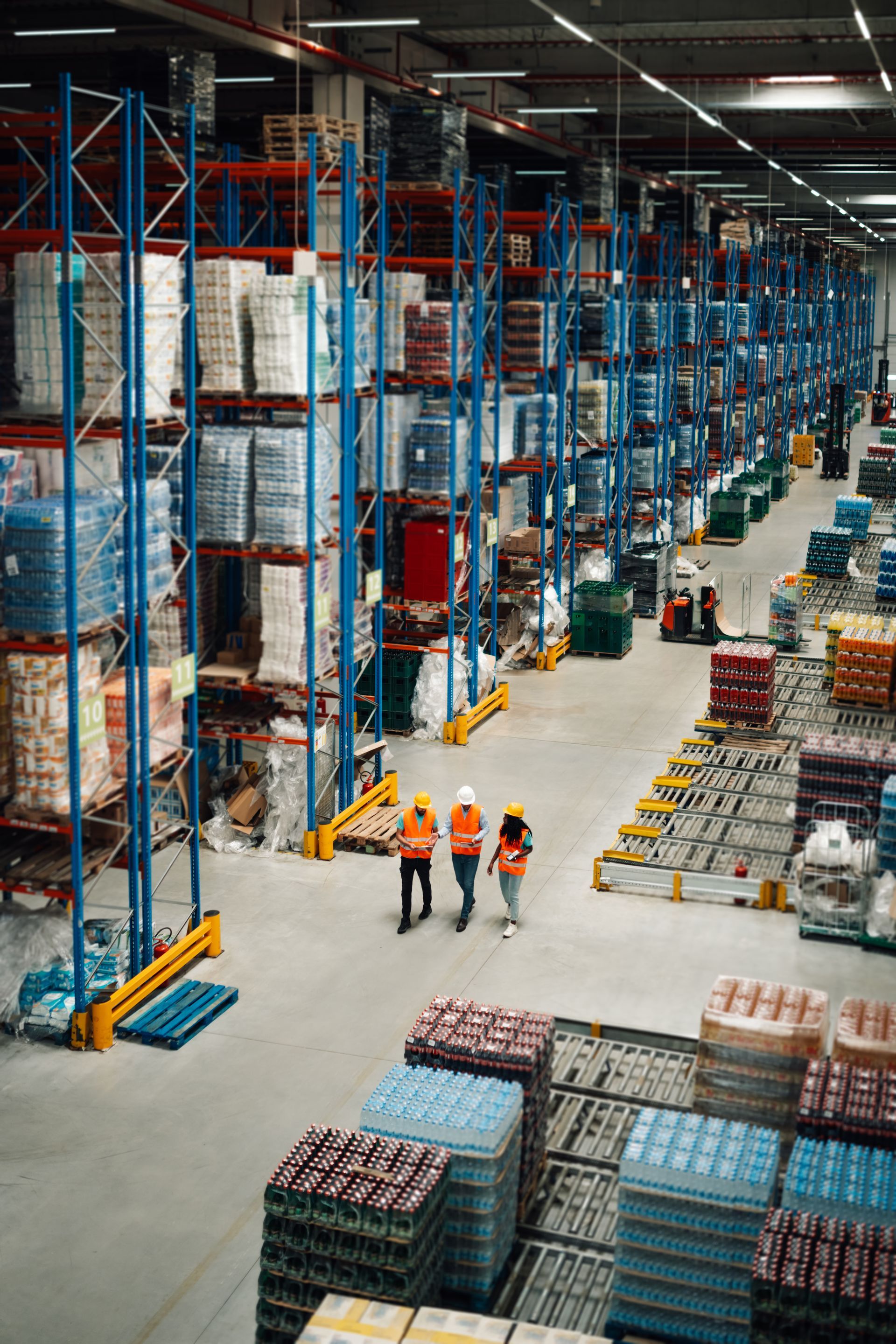 Warehouse interior with tall shelves stacked with goods. Three workers in safety vests walk along an aisle.