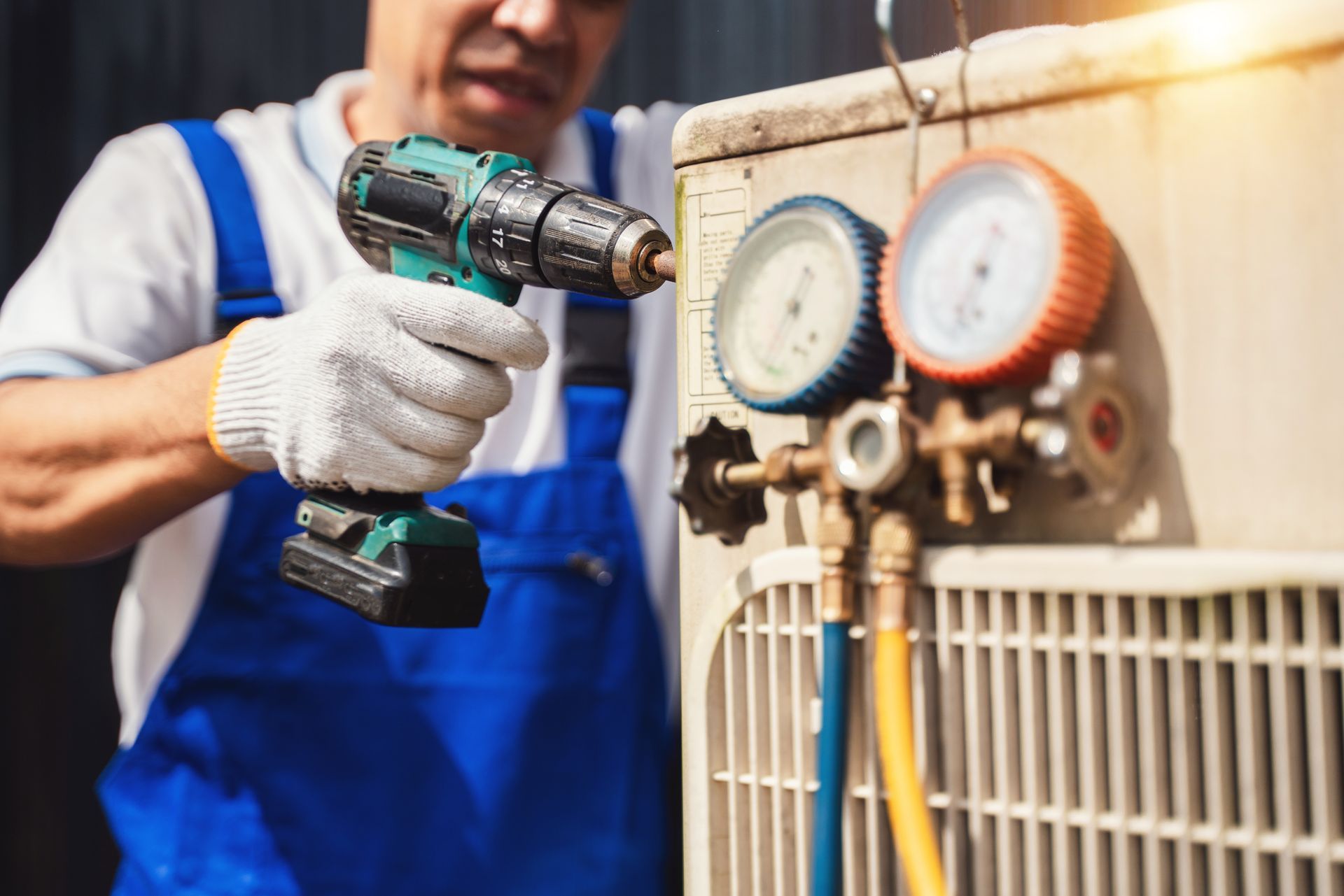 Technician in blue overalls using a cordless drill to service an HVAC unit with gauges attached.