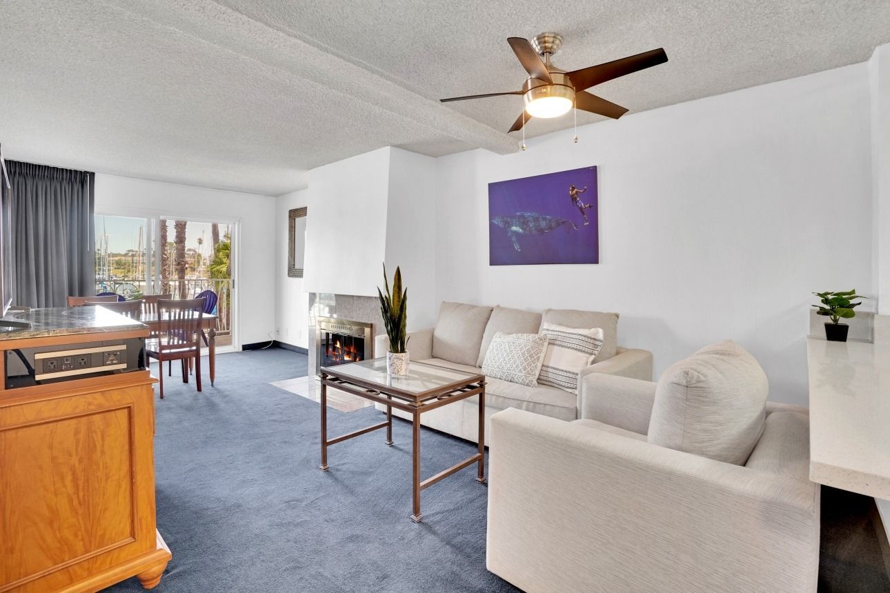 A living room with blue carpet, a beige sofa and armchair, a coffee table, a ceiling fan, and a view of a dining area.