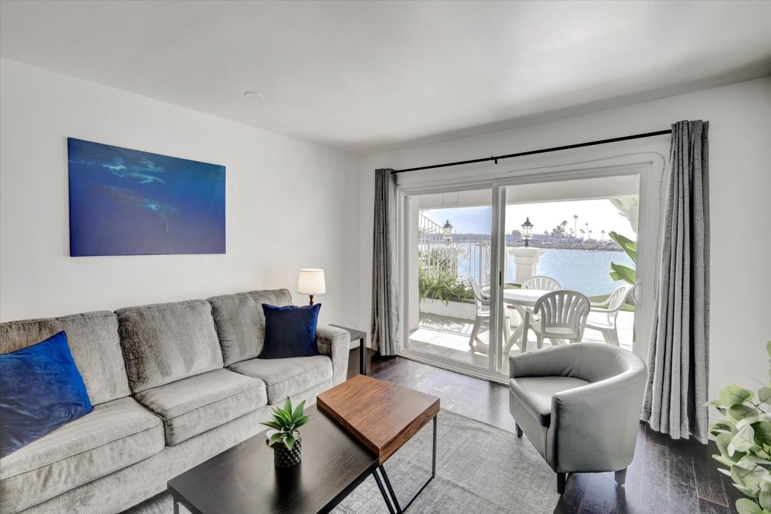 A living room with a gray sofa, blue pillows, a coffee table, and a sliding glass door opening to a patio by the water.