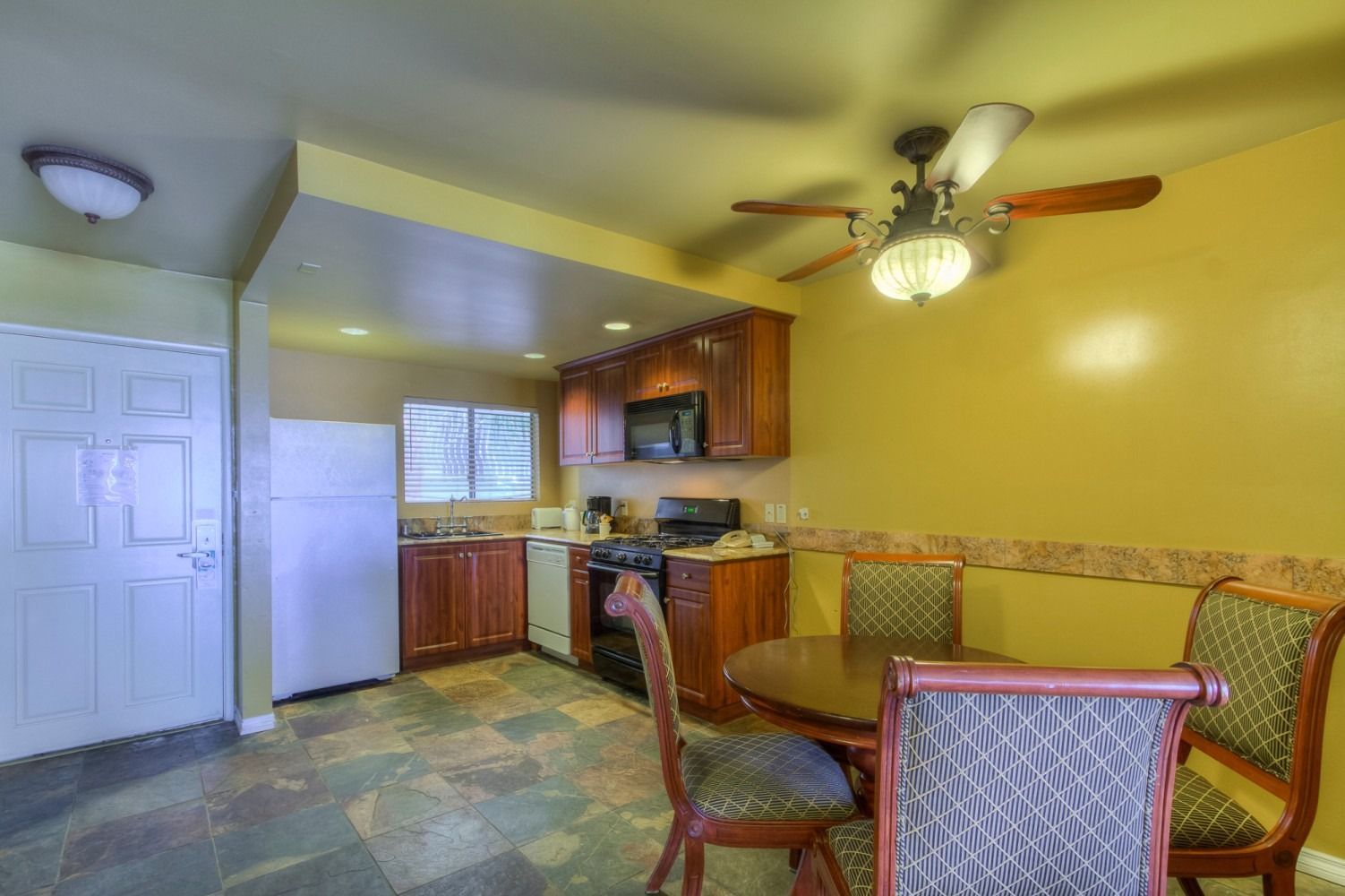 A kitchen with wood cabinets, yellow walls, and a dining set on stone-tiled flooring under a ceiling fan.