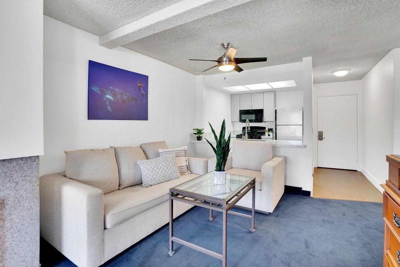 A furnished living room with a beige sofa, glass coffee table, and an open kitchen, set against a blue carpet.