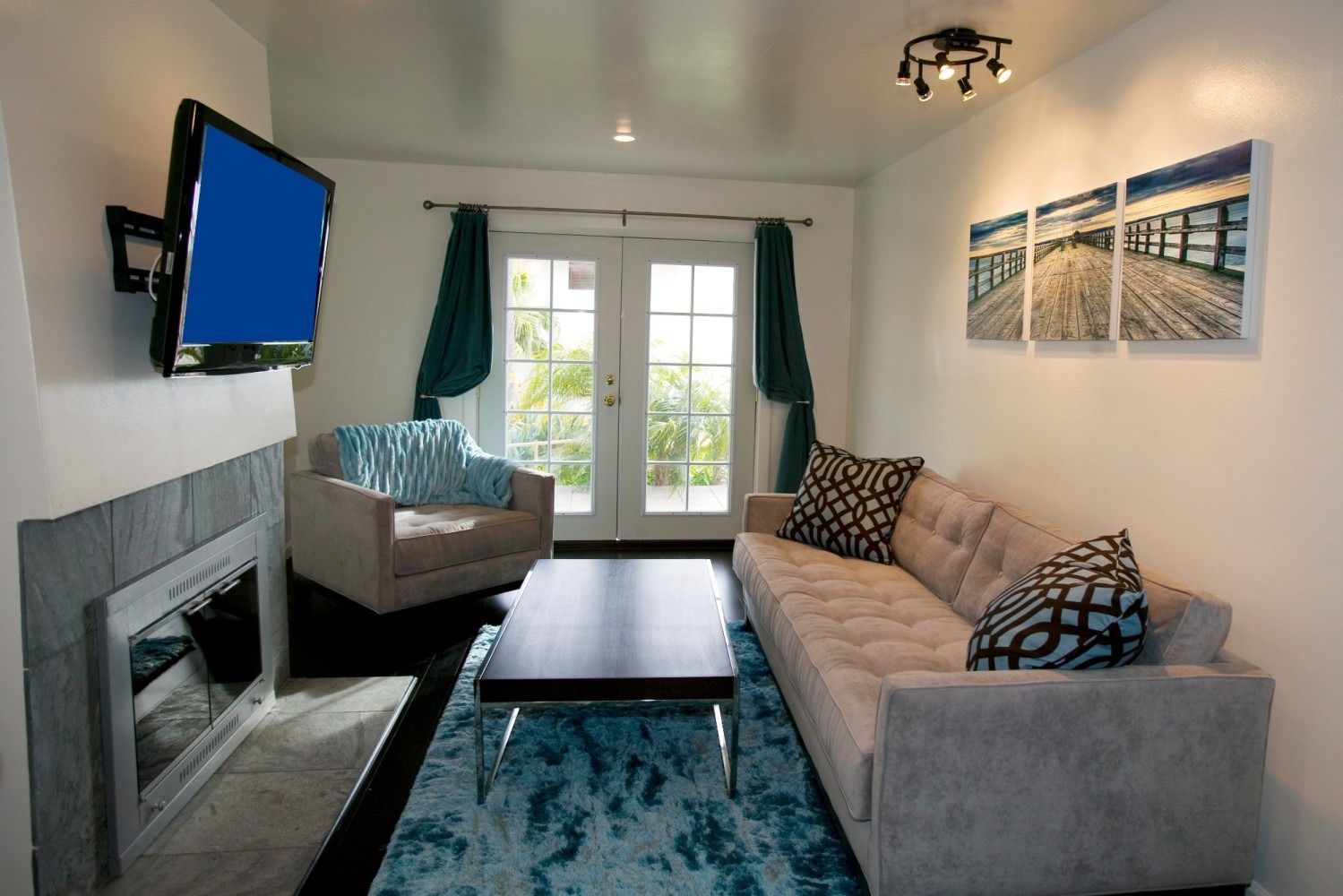 A cozy living room with a fireplace, TV, beige sofa, gray armchair, and a teal rug in front of French doors.