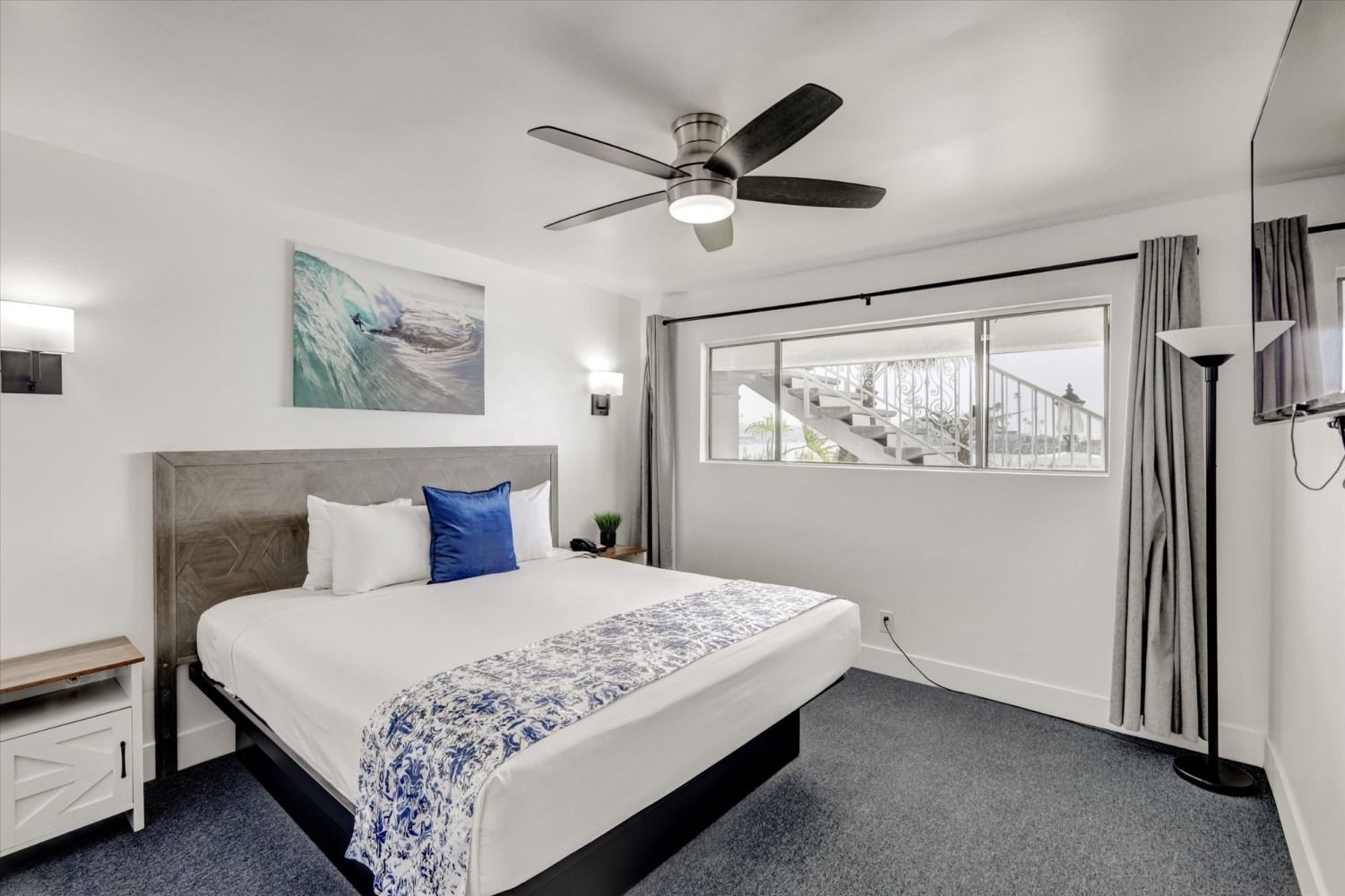 A bedroom with a king-sized bed, white walls, a ceiling fan, a blue-and-white art piece, and grey carpeting.