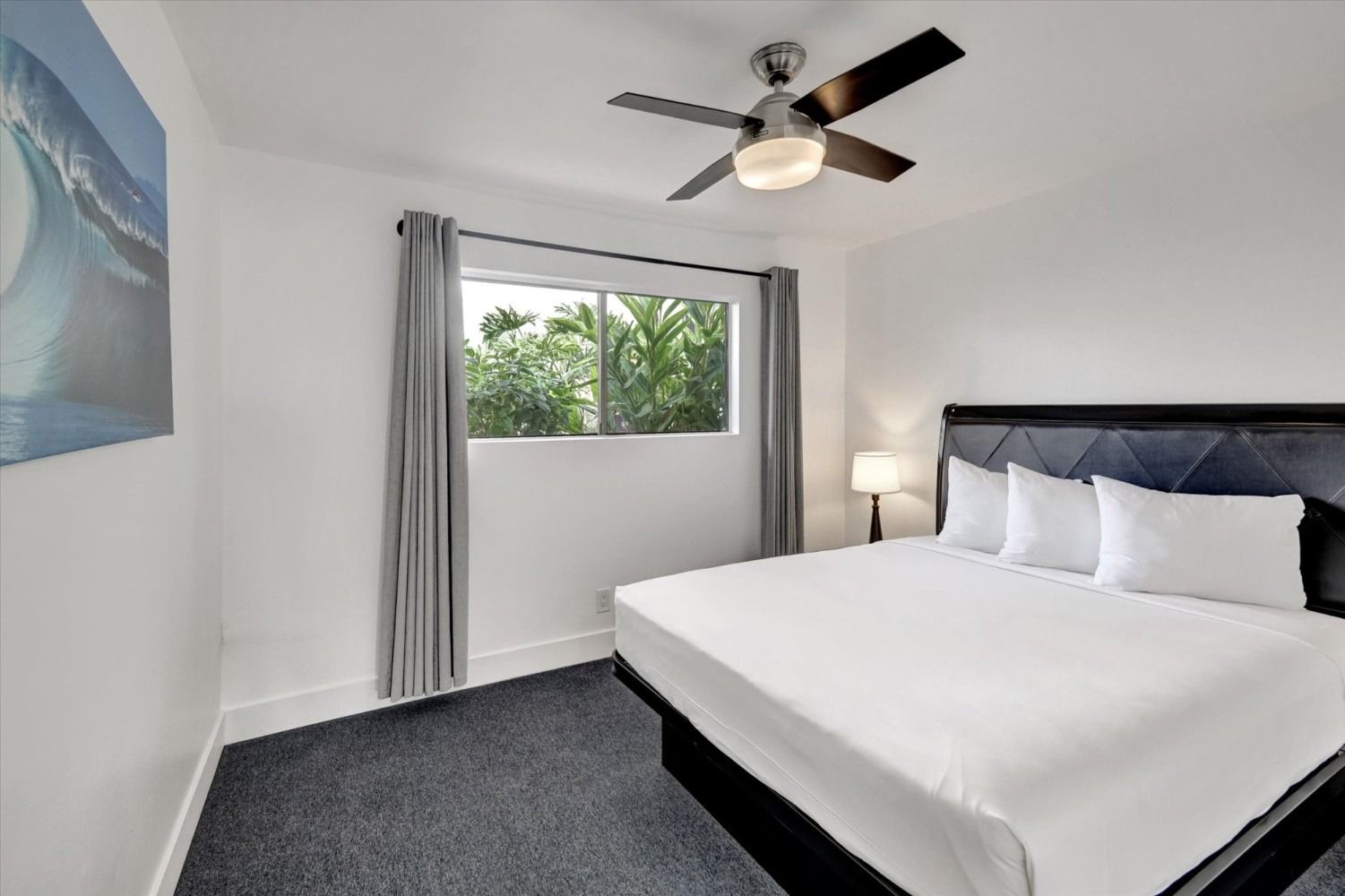 A bedroom with a king-sized bed, dark carpet, white walls, a ceiling fan, and a window with gray curtains.