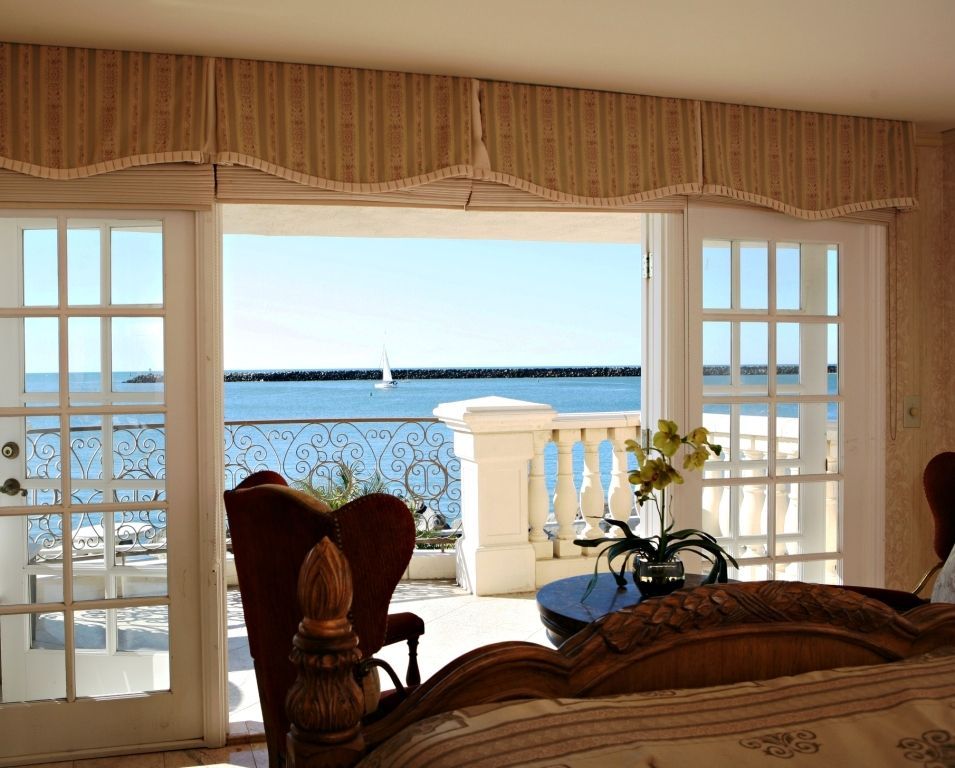 Bedroom with open French doors onto a balcony overlooking a blue ocean; a sailboat is visible.