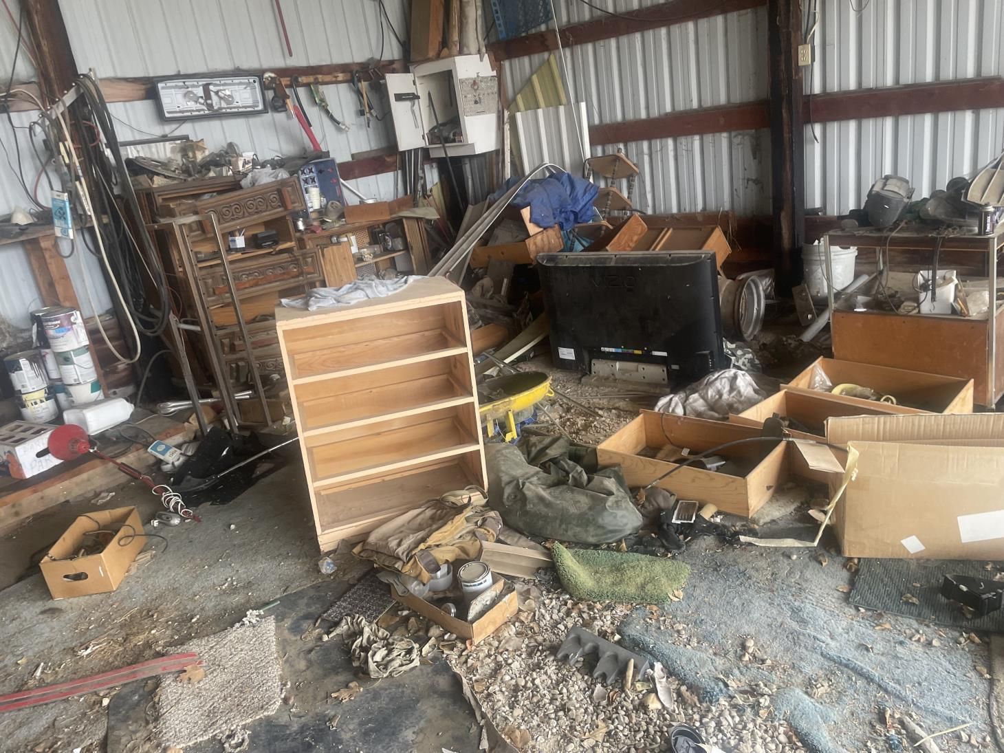 A cluttered workspace inside a shed featuring shelves, cardboard boxes, various tools, and equipment on a dusty floor.